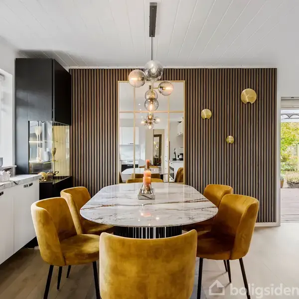 Marmorbord omgivet af gule stole står i spisestue med moderne lysekroner. Til venstre ses et køkken med hvide og sorte elementer. Glasdøre åbner ud til terrasse med haveudsigt.