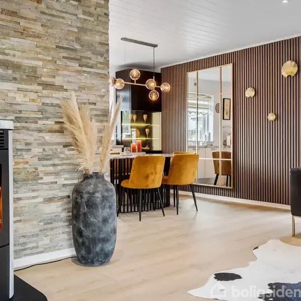 En moderne stue med en væghængt brændeovn i sort, der udstråler varme. En stor vase med dekorative tørrede græsser står ved siden af. Der er en spiseplads med gule stole og en spejlvæg i baggrunden.