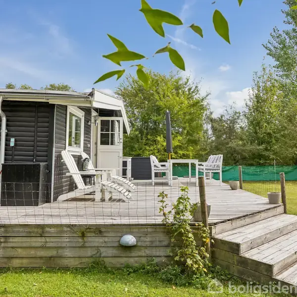 Et sort træhus med en veranda, der har hvide stole og et bord. Verandaen fører til en græsplæne, omgivet af træer og buske i en have.
