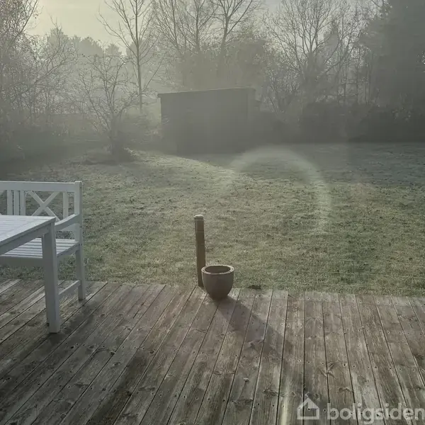 En hvid træbænk og bord står stille på en solbeskinnet træterrasse. I baggrunden ses en dugvåd græsplæne og tåget skovkrans.