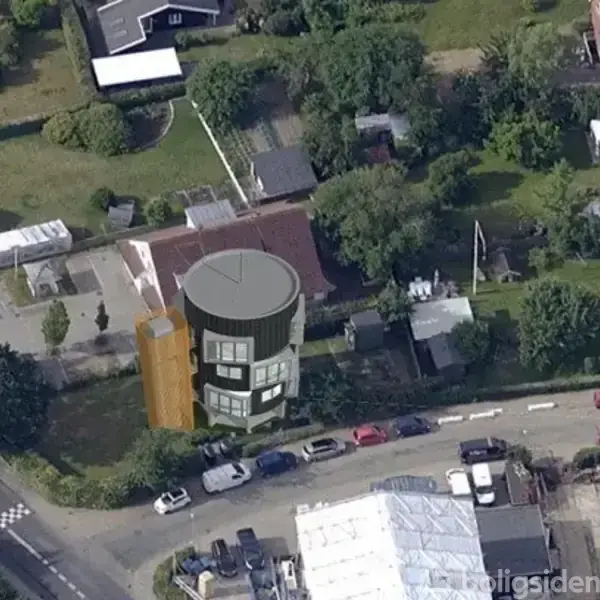 En stor bygning vises i en åben have ved en villavej med flere parkerede biler. Omkringliggende huse og træer skaber et tæt beboelsesområde.