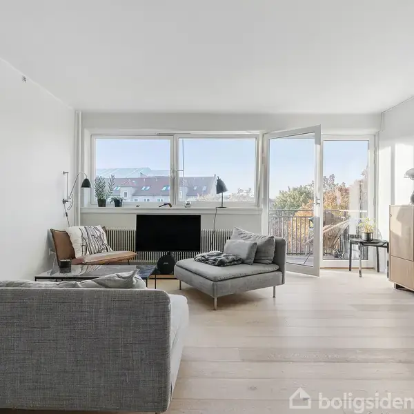 Sofa og lænestol arrangeret i en lys stue. I baggrunden er der store vinduer og en dør, der fører til en balkon. Lyse træmøbler og gulv fuldender rummet.