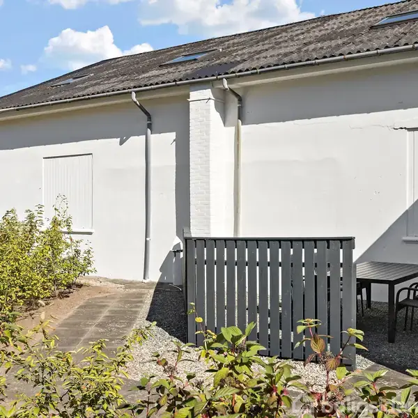 Bygning med hvid facade har et hegn foran, hvor et bord med tre stole er placeret. Omkring er der lav vegetation, og himlen er blå med få skyer.