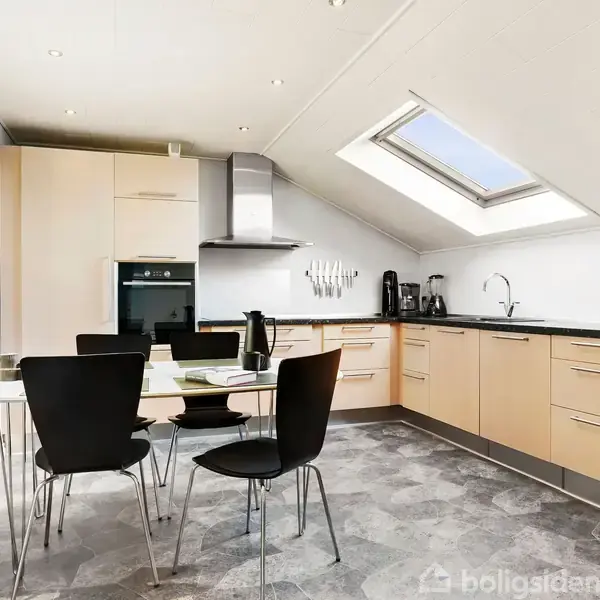 Et moderne køkken indeholder et spisebord med sorte stole. Køkkenet har lyse træskabe, indbygget ovn og en emhætte. Et ovenlysvindue giver naturligt lys til rummet.