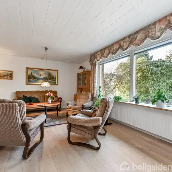 Stue med polstrede stole omkring et sofabord, lamper og malerier på væggene. Store vinduer med udsigt til en frodig have. Grønne planter dekorerer rummet.