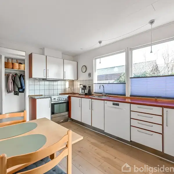 Køkken med hvide skabe, træbordplade og komfur i midten. Køkkenet har en spiseplads med lys træbord og stole. Vinduer til højre giver udsigt til omgivende huse og have.