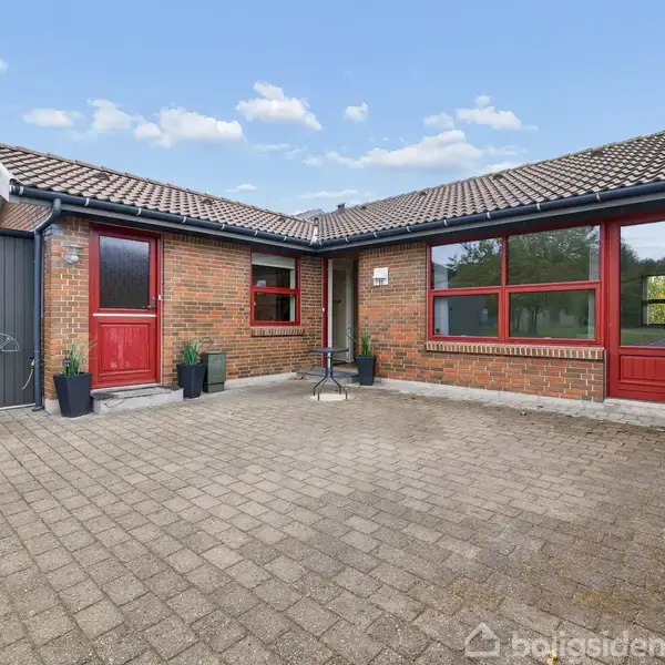 Rødt murstenshus med store vinduer og to røde døre, omkranser en flisebelagt gårdhave. Planter i potter står langs væggene under en blå himmel.