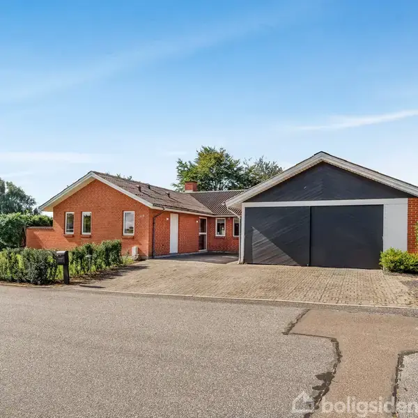 Eenfamiliehus i mursten står foran en dobbeltgarage. Husets indgang vender mod vejen, omgivet af velplejede buske og træer under en blå himmel.