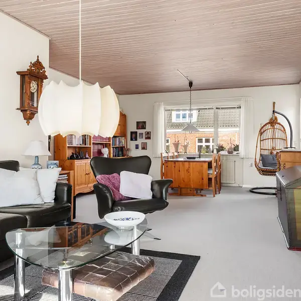 Stue med sorte lædersofaer set fra siden, moderne pendellampe og maleri på væggen. I baggrunden ses en bogreol, spisebord med stole, og et ophængt gyngesæde ved et vindue.