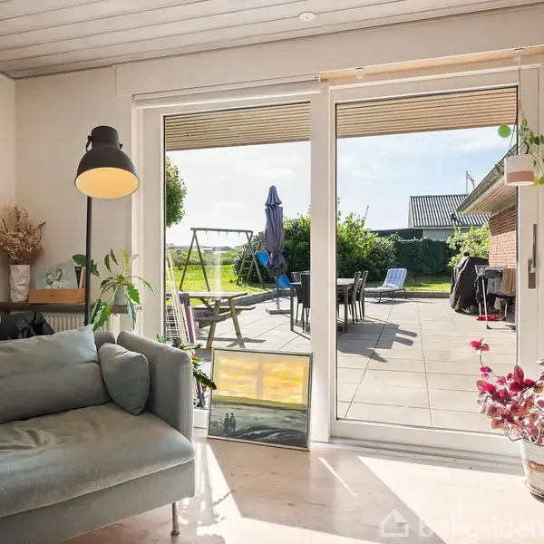 Stue med grøn sofa stående mod vindue; en gulvlampe tændt ved siden af; glasdøre åbner til solrig terrasse med havemøbler og udsigt til græsplæne og hus; planter omkring.