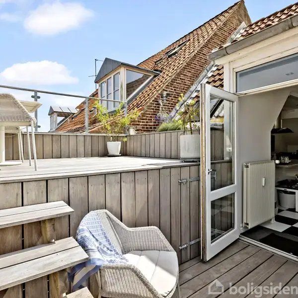 En terrasse med et hvidt cafébord og stole står hævet på træplatform. En åben terrassedør fører til et køkken med sort-hvidt ternet gulv. Sollys falder på terrassen.