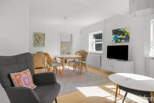 En stue med et gråt lænestol og et lille bord i forgrunden. Et spisebord med stole bagved. Væggene er dekoreret med malerier. Rummet er lyst og moderne.