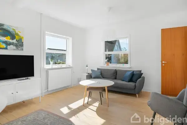 Gråt sofaarrangement står i en lys stue ved siden af et rundt hvidt sofabord. Fladskærms-TV placeret på hvidt skab. Maleri og tæppe tilføjer farve.