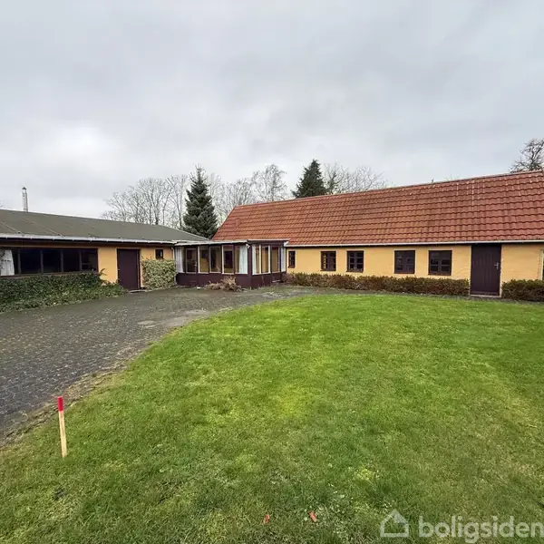 To bygninger med rødt tegltag og gule vægge står på en flisebelagt gårdsplads omgivet af en grøn græsplæne og træer.