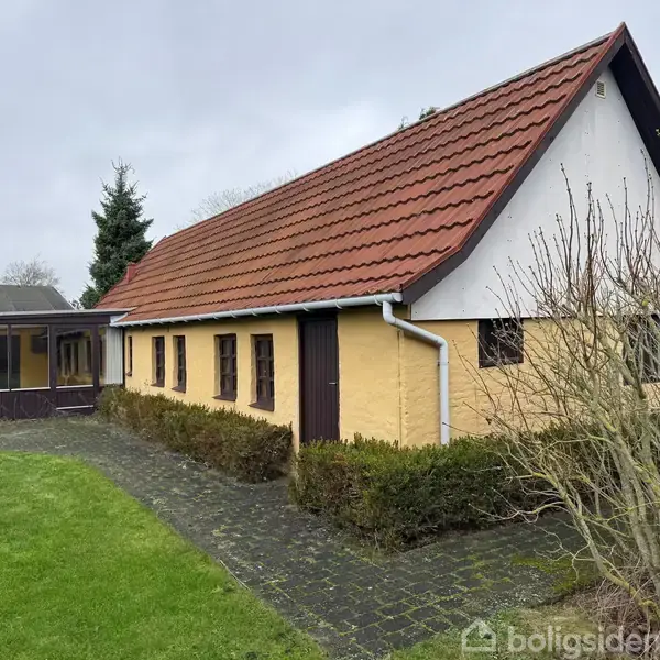 Gult landhus med rødt tegltag står i en have med græsplæne og brostensbelagt sti. Buske grænser op til huset, der har flere vinduer langs siden.