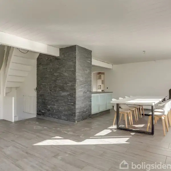 Spisestue med lang hvid spisebord og stole, der står nær en mørk stenvæg; til venstre er en hvid trappe. Værelset er lyst og rummeligt med store vinduer.