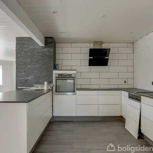 Et moderne køkken med hvide skabe og grå bordplader. En ovn er indbygget i væggen under en sort emhætte. Rummet har store vinduer og en stenvæg til venstre.