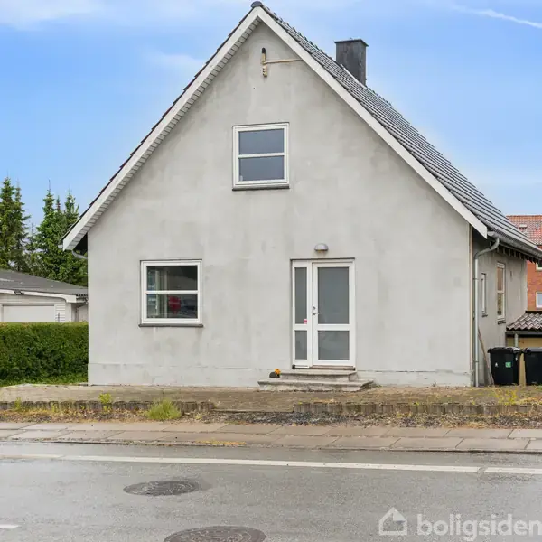 Et hvidt hus med skråt tag står roligt ved en vej. Omkring huset er en lav hæk, og et træ står til venstre. Vejret ser klart ud.