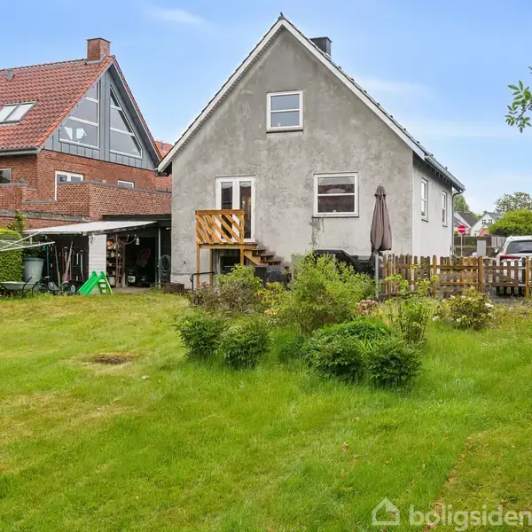 Et gråmalet hus med skråtag står i en baghave. Træterrasse med trappe og parasol. Omgivet af grøn græsplæne og buske. Nabohuset er rødt murværk.