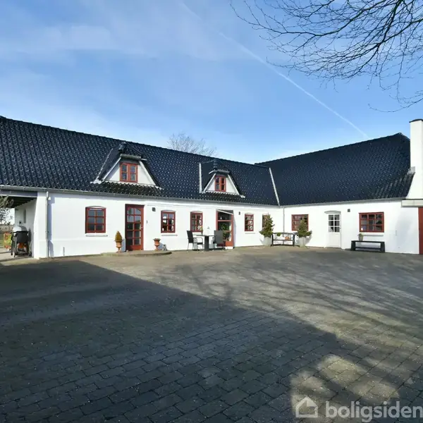 Et hvidt bondehus med sort tag står stille i en gårdsplads. Bygningen har flere røde vinduer og en dør. Der er sollys og træer i baggrunden.