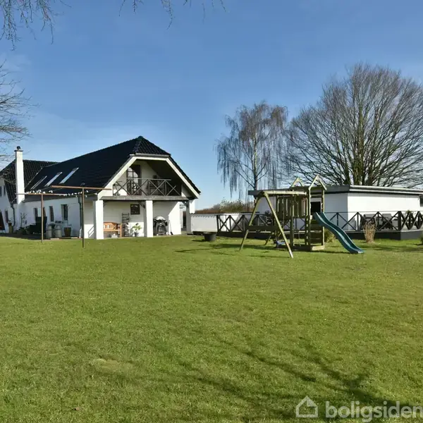 Hus i hvid facadestil med sort tag står på en stor, grøn græsplæne. Ved siden af er en legeplads med gynger og rutsjebane. Træer omkranser haven.