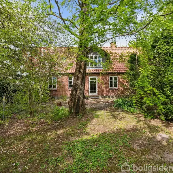 Et gammelt murstenshus med rødt tegltag står blandt tætte, grønne træer og buske. Haven er vildtvoksende og frodig, skaber en rolig og naturlig atmosfære omkring huset.
