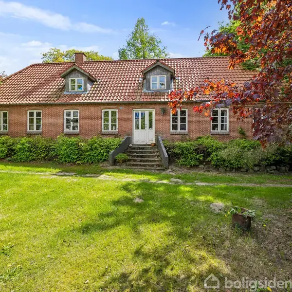 En rød murstensvilla med rødt tegltag står foran en velplejet græsplæne og omgivet af frodige buske. To mindre vinduer er placeret i kvistene på taget.