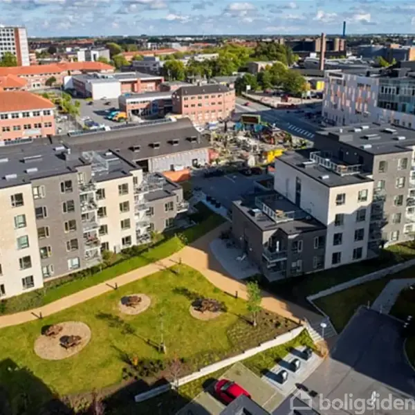 Flere moderne lejlighedsbyggerier står tæt sammen, omringet af anlagte grønne arealer med stier og cirkelformede områder. Bybaggrund med yderligere bygninger og byens skyline ses i horisonten.