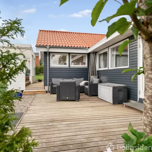 Hus med sort træbeklædning og rødt tag står på en terrasse med havemøbler. Grønt løv og grusgang omkranser terrassen, skaber en hyggelig udendørs atmosfære.