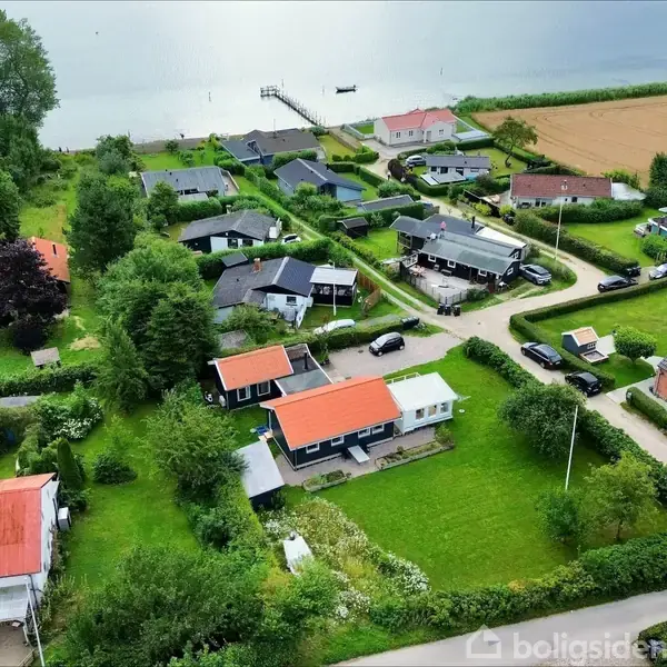 Et fugleperspektiv viser en række huse med haver, der grænser op til en vej. Havet og en bådbro er synlige i baggrunden, omgivet af marker og træer.