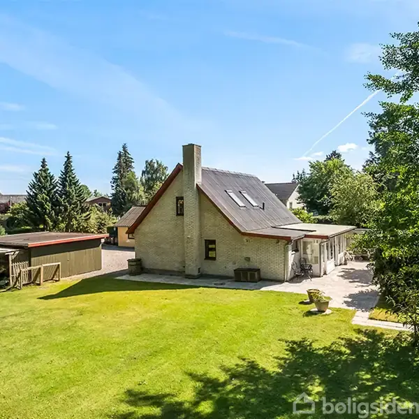 Et hvidt hus med skråt tag og skorsten står i en grøn have med træer og buske. Skyfri blå himmel og andre huse i baggrunden.