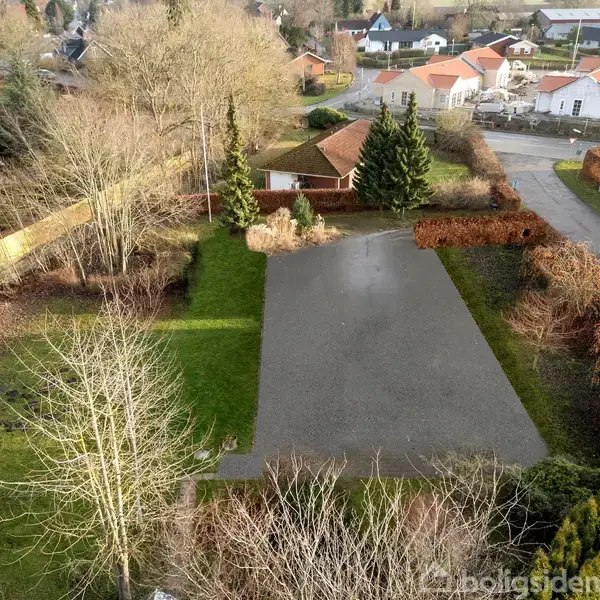 En tom grund ligger stille omgivet af træer og buske, med stier der leder til et nabohus og en vej. I baggrunden ses flere huse i et villakvarter.