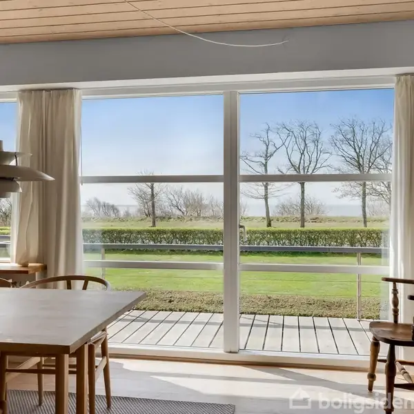 Spisebord med stole står foran store vinduer, der viser en udsigt til en grønt område med træer. Rummet er lyst indrettet med hvide gardiner og en hængende lampe.