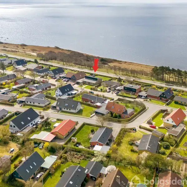 Luftfoto af en beboelsesområde med huse omkring veje. Rødpil peger på et specifikt hus nær kysten med havet i baggrunden. Grønt område omgiver husene.