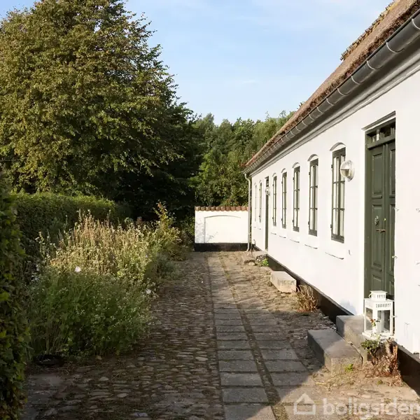 Hvidt hus med mørkegrønne døre og vinduer står ved siden af en brostensbelagt sti i en frodig have med høje buske og træer i baggrunden.