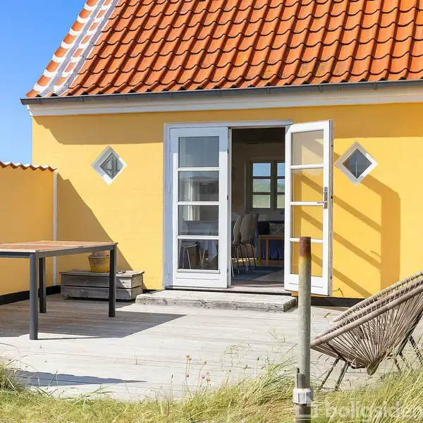 Gulmalet hus med rødt tegltag og terrassedør åben til en solrig terrasse. Hængekøje og bord står udendørs på træplanker omgivet af græs.