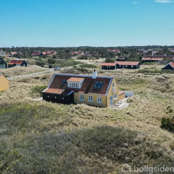 Hus med gult murværk og rødt tag står alene i et åbent klitlandskab. I baggrunden ses flere spredte sommerhuse omgivet af sand og vegetation under en klar blå himmel.