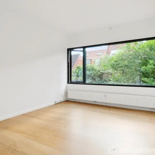 Et lyst værelse med trægulv. Store vinduer tilbyder udsigt til træer og bygninger udenfor. Vægge og loft er hvide, og der er en radiator under vinduet.