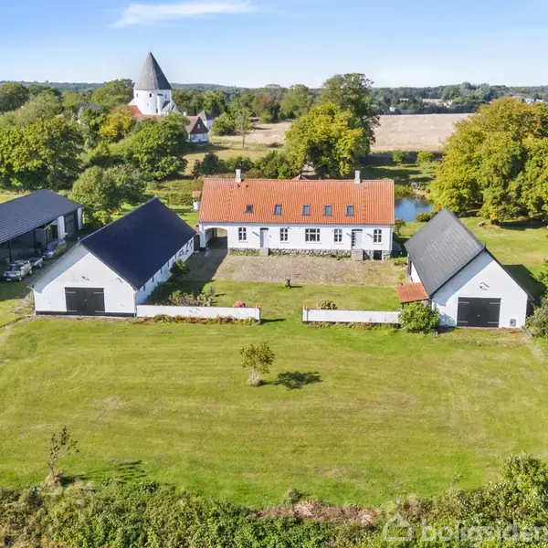 En hvid gård med rødt tag omgivet af flere bygninger står på en stor grøn mark. I baggrunden ses en kirke med et hvidt tårn og træer.