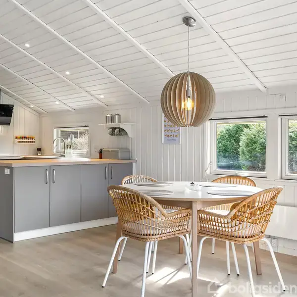 Et rundt bord med flettede stole står i et lyst køkken-alrum. Køkkenet har grå skabe, vinduer med udsigt til grønne træer og en dekorativ lampe hænger over bordet.
