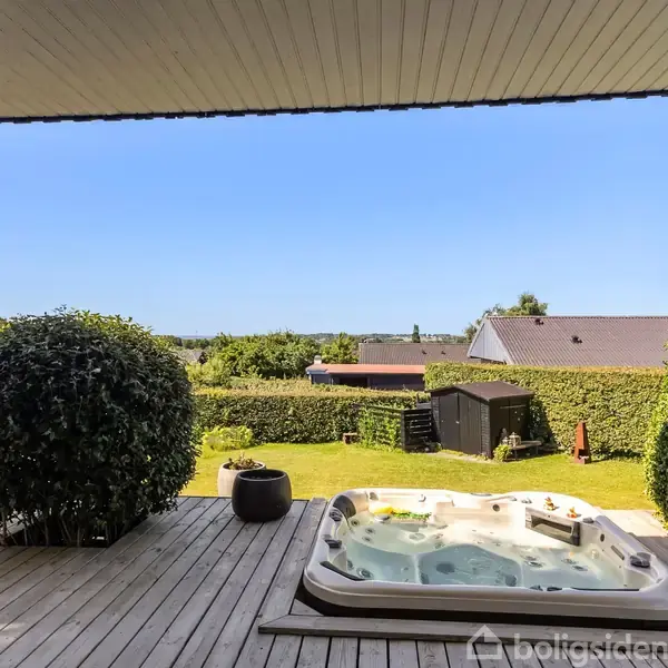 Boblebad står på en træterrasse og er omringet af grønne buske og græsplæne i en have. Baggrunden viser udsigt over naboens hustag og en klar blå himmel.