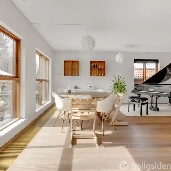 Et moderne rum med et stort flygel, trægulve og spisebord med stole. Store vinduer lader naturligt lys strømme ind. Planter og indbyggede hylder skaber en indbydende atmosfære.