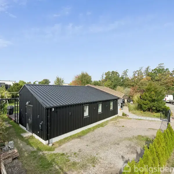 Sort étplanshus står stille på en grusbelagt grund i forstadsområde med grusvej til venstre og buskads i baggrunden. Klar himmel giver en lys atmosfære.