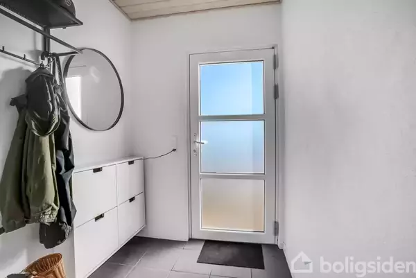 En entré med en glasdør, der lader uskarpt lys komme ind. Jakker hænger på en knagerække ved siden af et rundt spejl. Skoskabe står langs væggen.