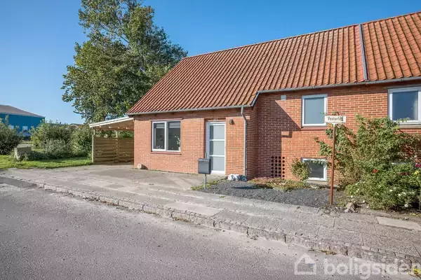 Rødt murstenshus med tegltag har en indgang og to store vinduer. Skiltet siger "Vesterdal". Det omgives af en betonsti og nogle træer i baggrunden.