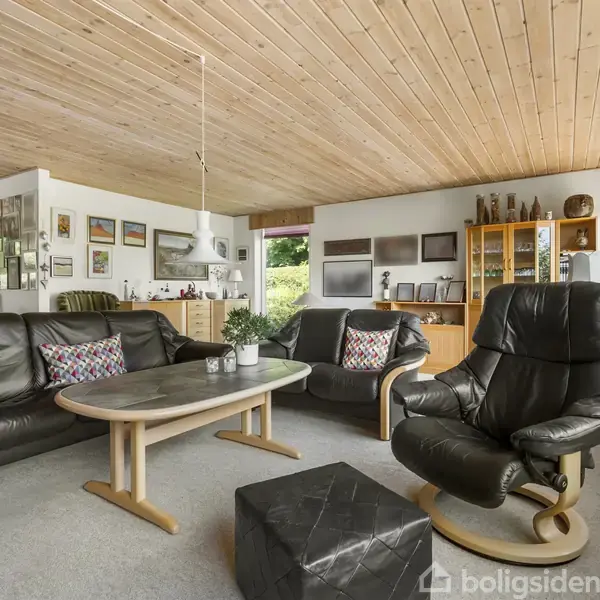 Sort lædersofa og lænestol står i en stue med træloft, hvor et sofabord er placeret foran. Omkring er der malerier på væggene og et vitrineskab.
