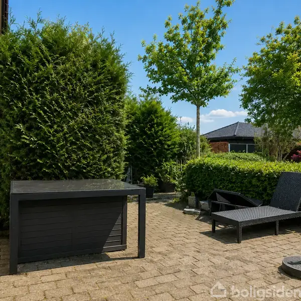 Et sort havebord står på en flisebelagt terrasse, omgivet af grønne træer og buske. En sort liggestol er placeret ved siden af, under en blå himmel.