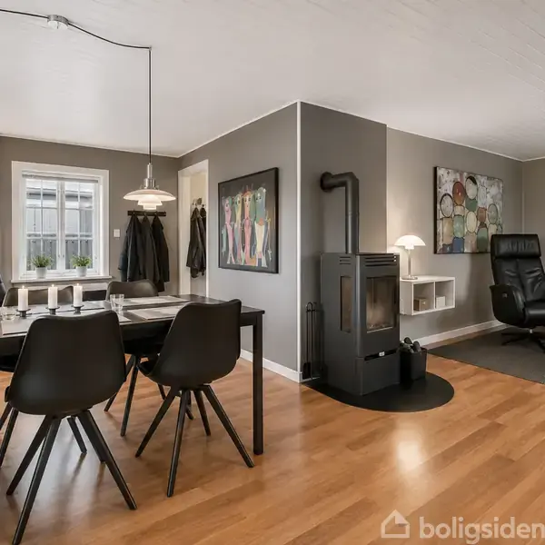 Spisebord med sorte stole står i et lyst, moderne rum. Ved siden af er en brændeovn, og i baggrunden ses en lænestol og sofa. Væggene har billeder. Ingen tekst.