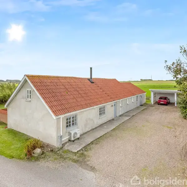 Et hvidt bondehus med rødt tegltag står stille ved en grusvej. Ved siden af huset ses en carport med en rød bil, omgivet af åbent landskab og blå himmel.