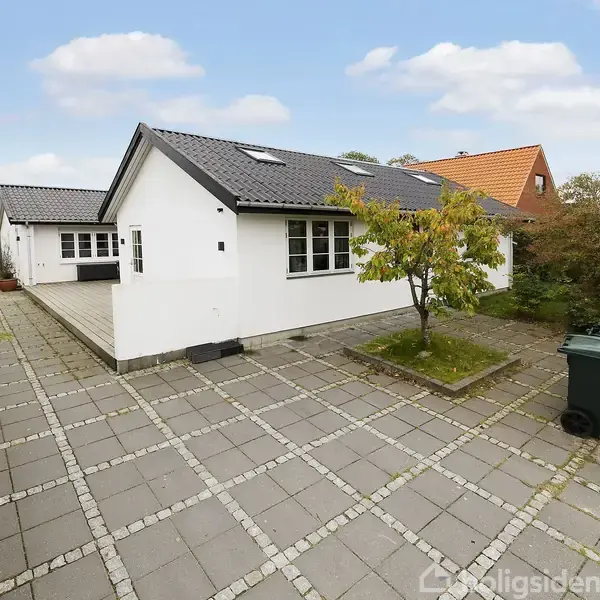 En hvid, én-etagers bygning med sort tag står stille i en have. Flisebelagt terrasse, omgivet af grønne buske og et enkelt træ. Senere ses en anden rød-orange bygning i baggrunden.